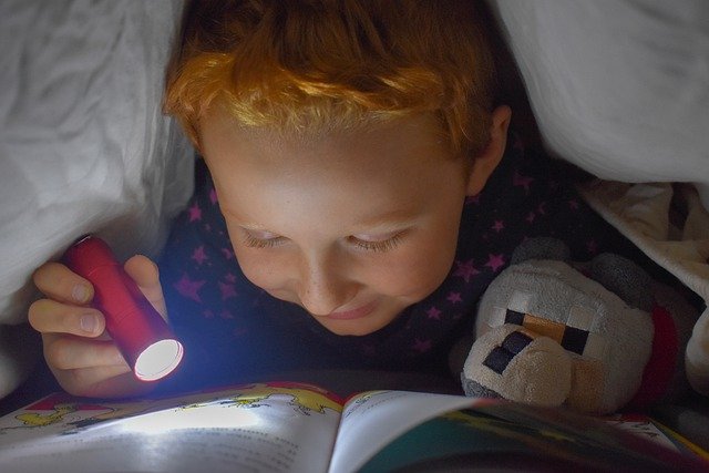 boy reading under bed covers with flashlight